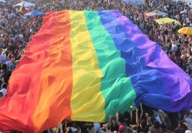 Uma multidão ergue uma bandeira com as cores o arco-íris gigante