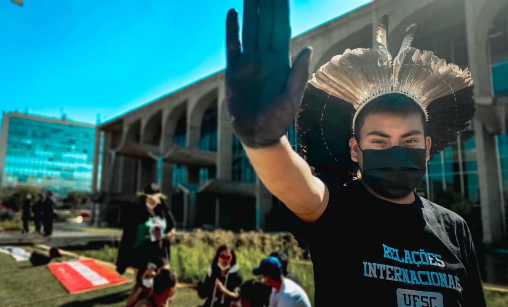 Jovem indígena de cocar de penas bege e pretas, máscara cirúrgica e camiseta do curso de Relações Internacionais da UFSC levanta a mão, pintada de preto, com a palma para a frente. Ao fundo, Ministério da Justiça.