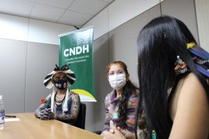 Dois estudantes com grafismos e adereços de penas conversam com promotora na sala do Conselho Nacional dos Direitos Humanos. Todos estão de máscara.