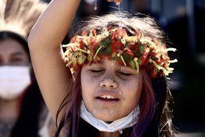 Criança indígena com adereço de flores na cabeça, máscara cirúrgica abaixada e pintura de jenipapo no rosto. Ela está falando e deixa pingar um líquido transparente de sua mão, acima da cabeça.