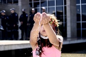 Criança com adereço florido e máscara abaixada usa as mãos em concha para jogar água à sua frente. Ao fundo, policiais montam guarda no Ministério da Justiça.