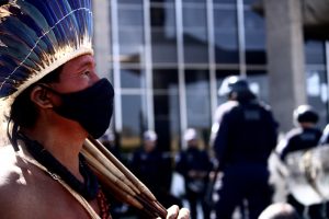 Em primeiro plano, homem indígena de cocar de penas azuis, colares e máscara de pano. Ao fundo, desfocados, prédio do Ministério da Justiça e policiais.