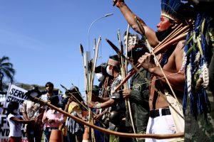 Parte das pessoas do protesto. A maioria é indígena e leva cocares, colares e arcos e flecha. Ao fundo, alguns seguram cartazes com frases de ordem.