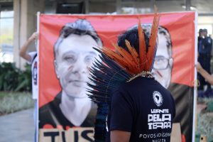 Homem indígena com cocar de penas vermelhas e azuis apoiado na nuca e camiseta do Levante pela Terra observa, em primeiro plano, cartaz que pede justiça pelas mortes de Dom Phillips e Bruno Pereira.