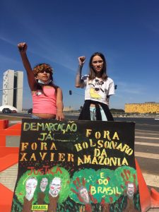 Duas crianças de punho erguido seguram cartazes pintados à mão: Demarcação já; Fora Xavier; Fora Bolsonaro da Amazônia e do Brasil. Nas árvores pintadas, estão as fotos de Dom e Bruno com pinturas indígenas no rosto.