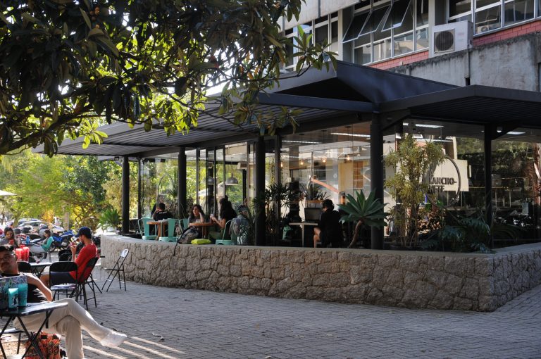 Lanchonete bem decorada ao ar livre, com plantas, parede de pedra, cadeiras de metal minimalistas e muitos clientes.