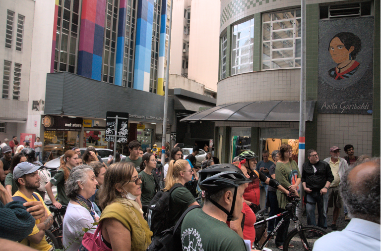 Rua em frente à Câmara repleta de manifestantes, muitos deles ciclistas utilizando capacete. Há um mural da Antonieta de Barros no prédio em frente.