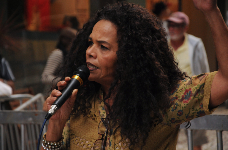 Mulher negra madura com expressão determinada, roupa colorida e braceletes discursa com um microfone.